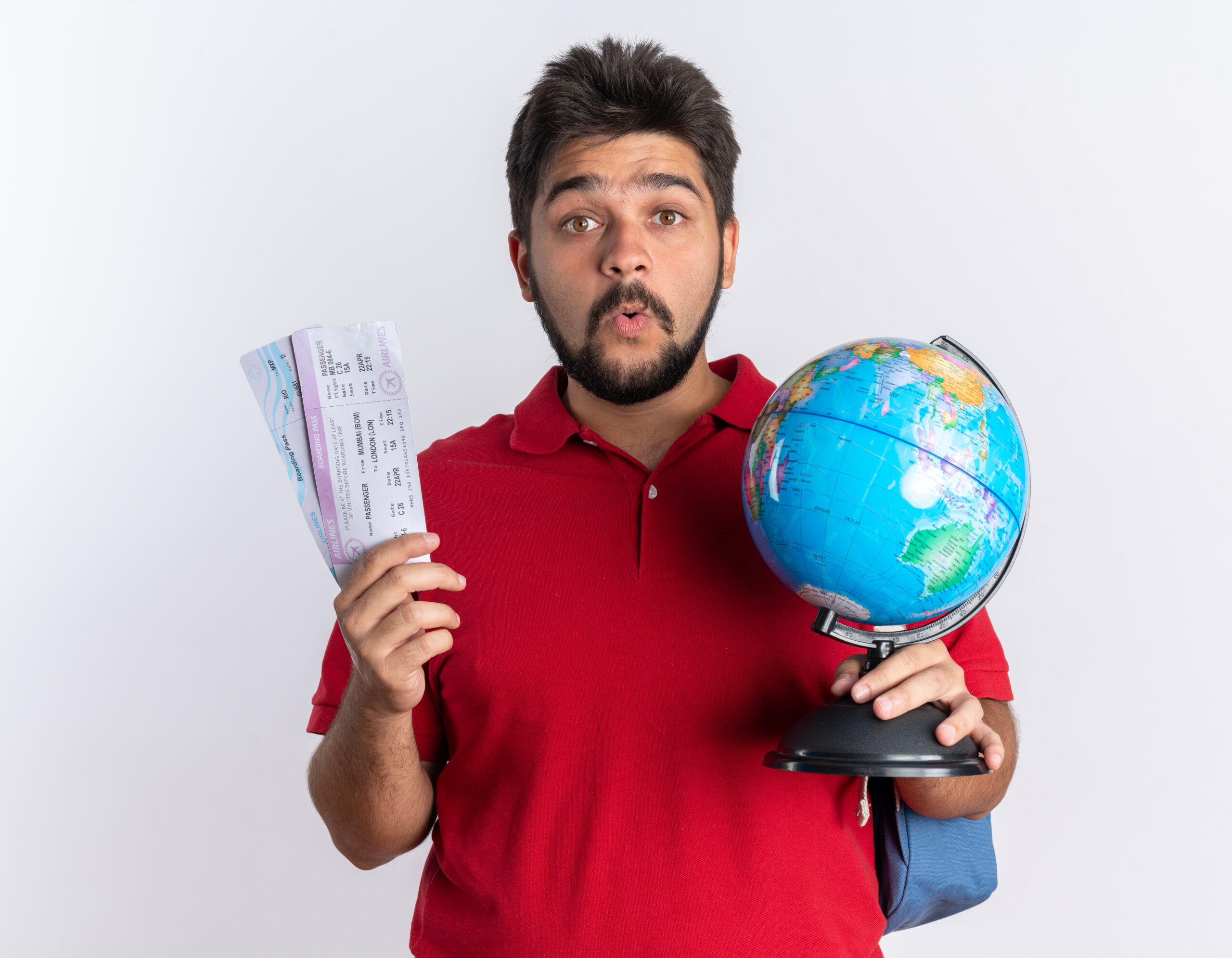 young bearded student guy in red polo shirt with backpack holding air tickets and globe looking at camera surprised standing over white background